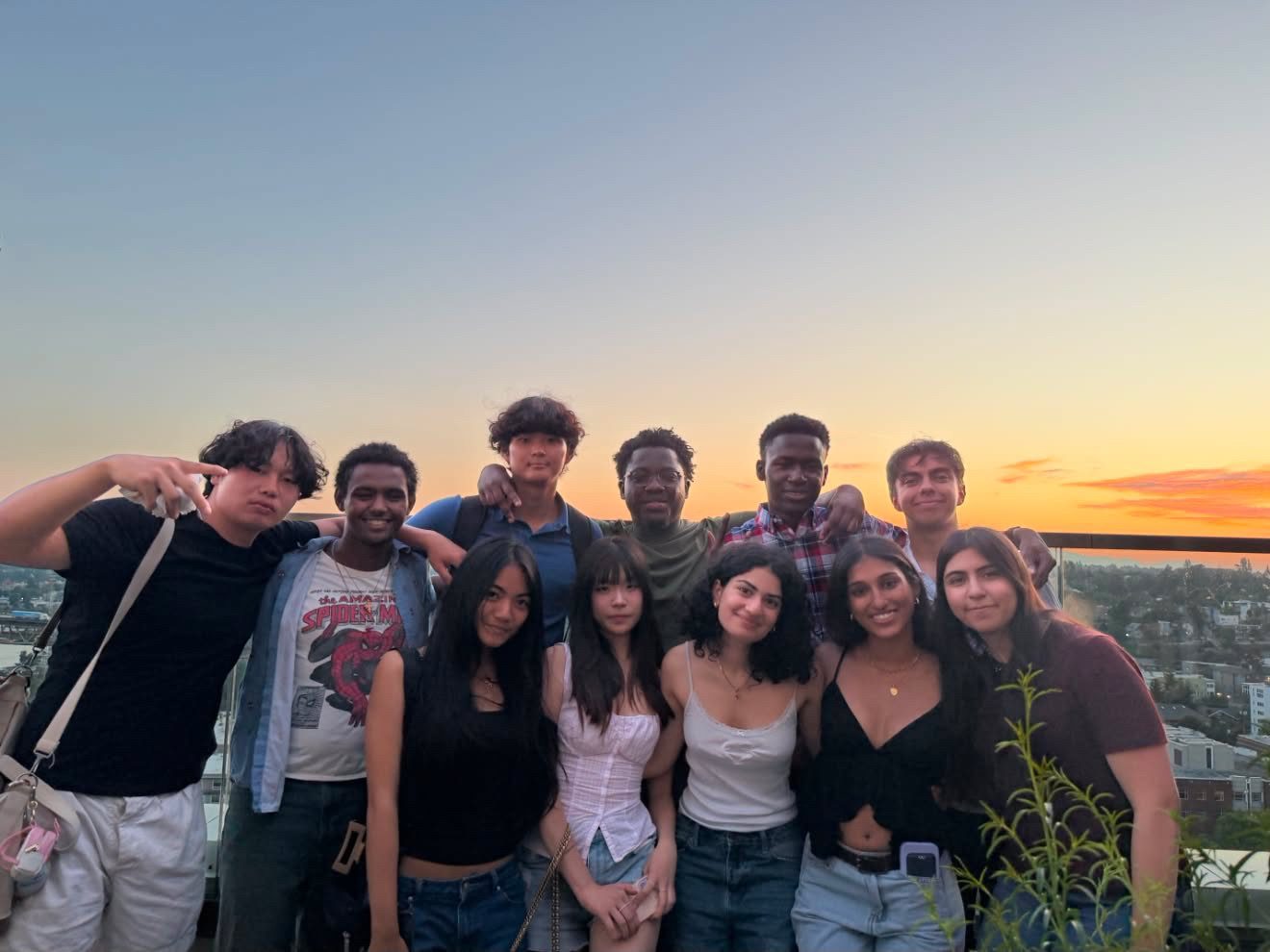 About a dozen Amazon Future Engineer scholars crowded together on a rooftop at sunset, arms around each other, a city skyline and orange sky behind them.