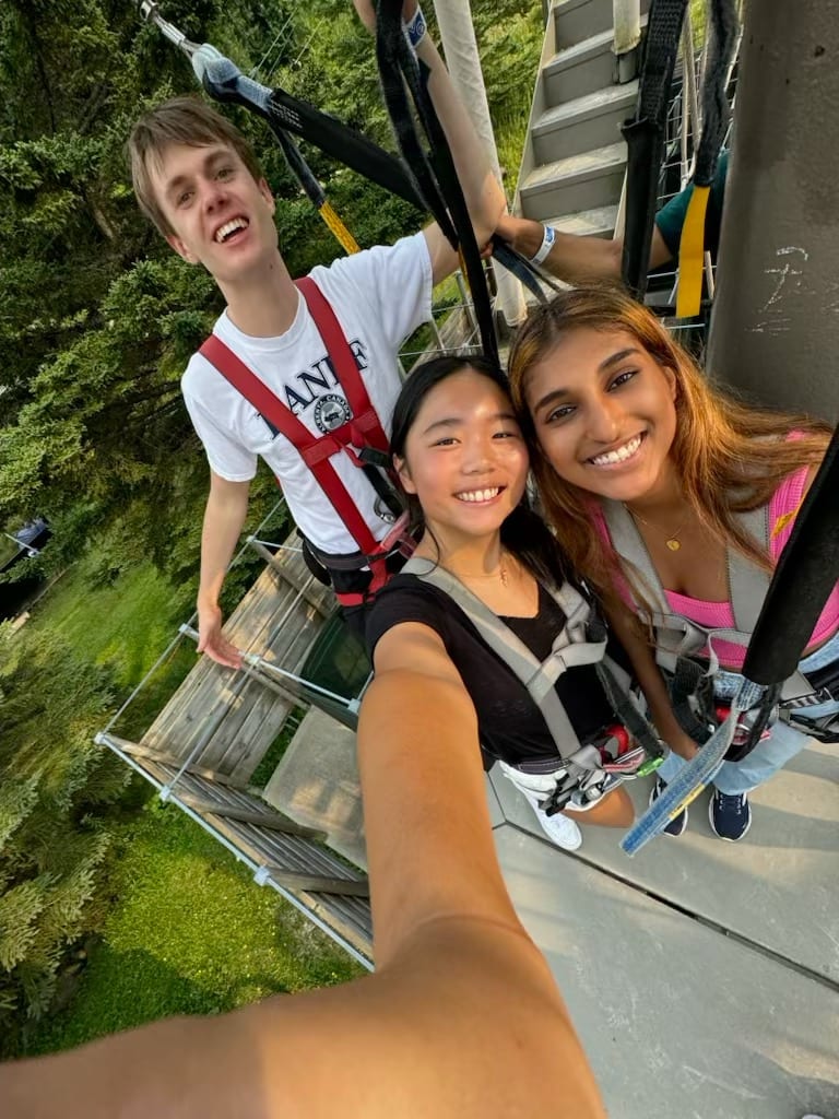 Selfie from the top of a zip-line tower — three Senators in climbing harnesses, treetops and grass far below.