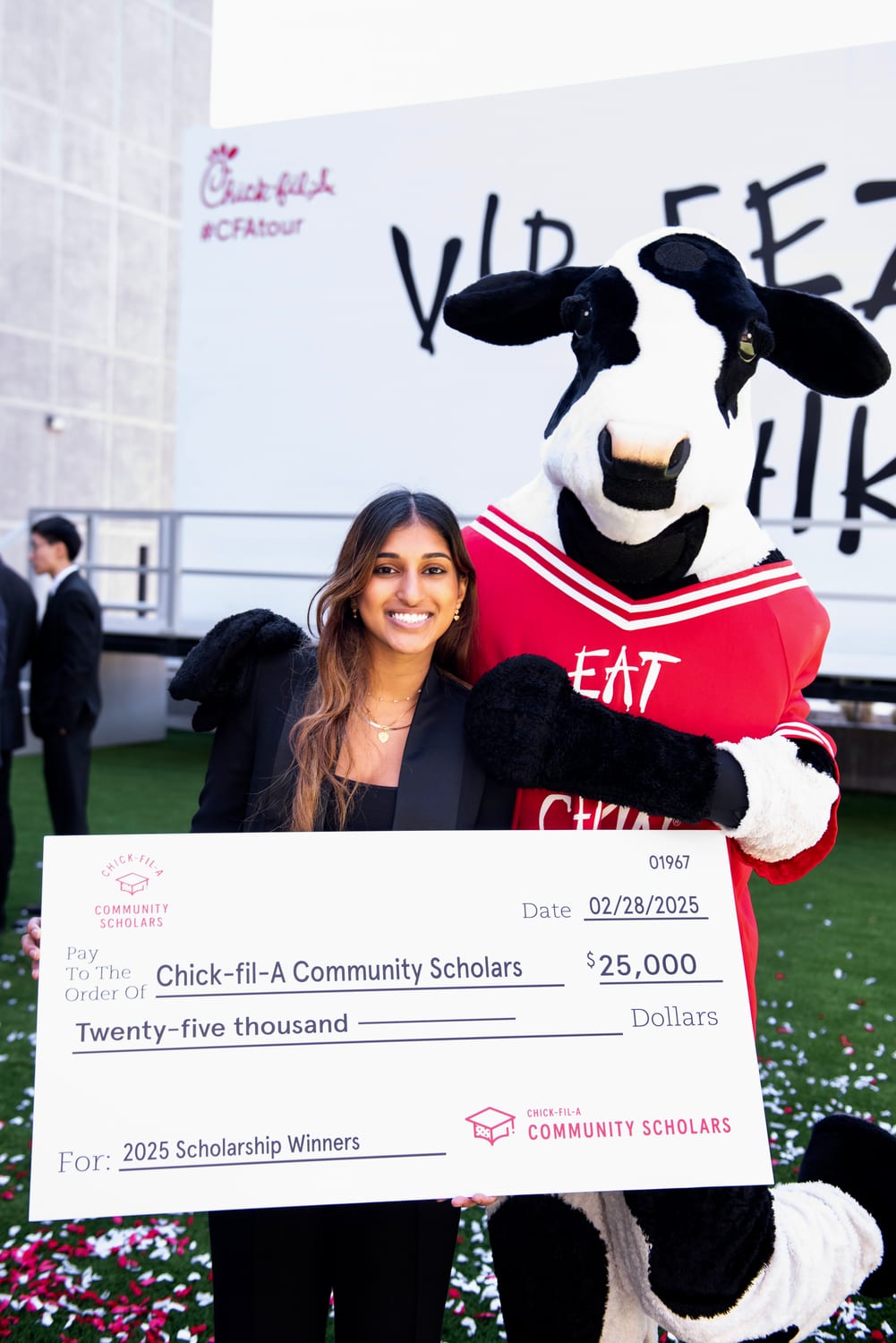 Ava in a black blazer holding an oversized Chick-fil-A Community Scholars check — $25,000, dated 02/28/2025, “For: 2025 Scholarship Winners” — beside the Chick-fil-A Cow mascot in its red “Eat Mor Chikin” jersey, confetti scattered on the turf.