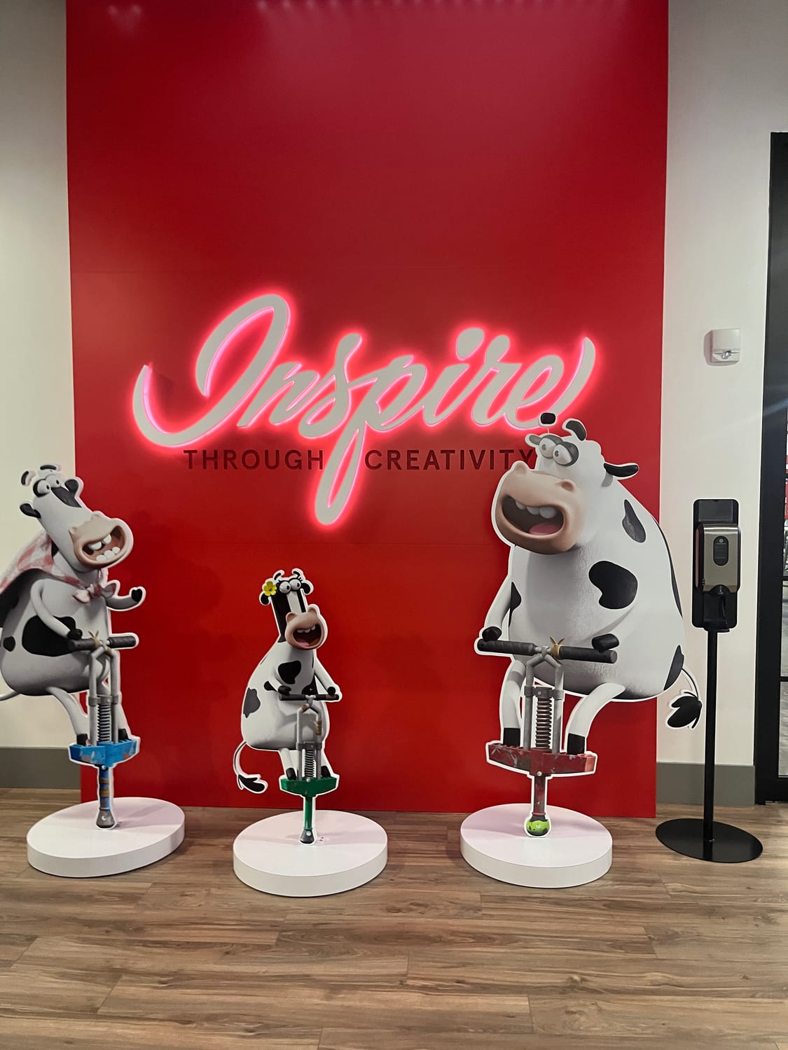 A Chick-fil-A Support Center display: a red wall with a neon-script “Inspire” over “Through Creativity”, fronted by three cartoon cows sculpted riding paint-tipped pogo sticks.