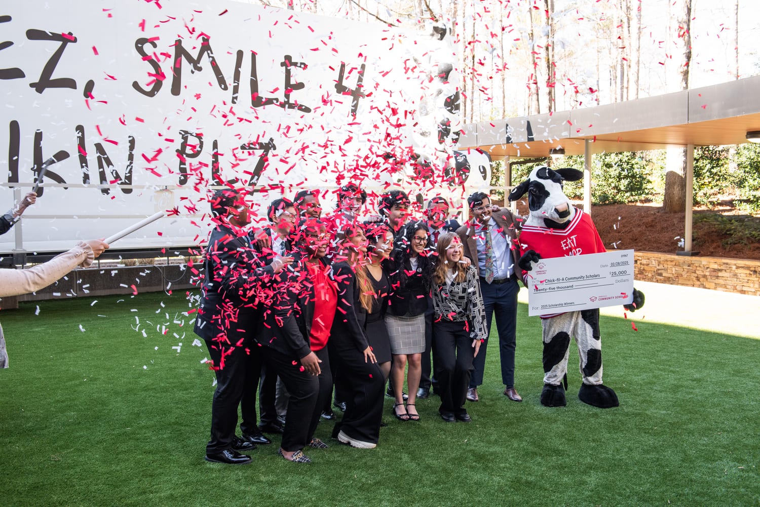 The scholarship cohort crowded together holding the giant $25,000 check beside the Cow mascot as red and white confetti cannons go off in front of the “Eat Mor Chikin” cow truck.