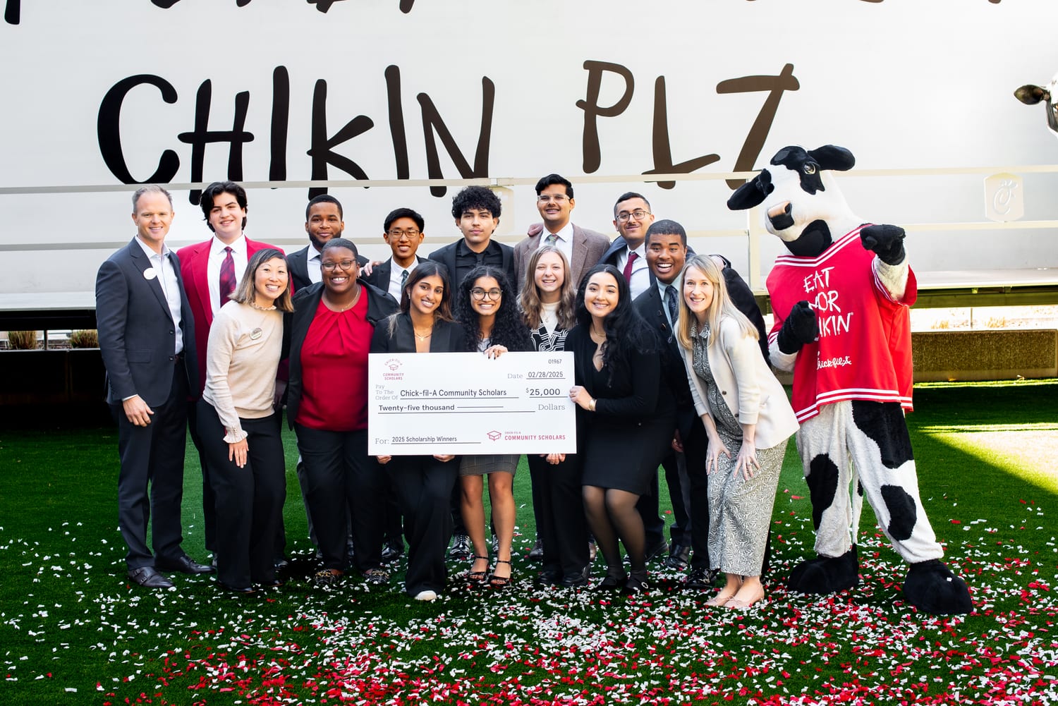 The full cohort of scholars with Chick-fil-A staff and the Cow mascot gathered around the $25,000 check on confetti-covered grass in front of the “Chikin Plz” cow truck.