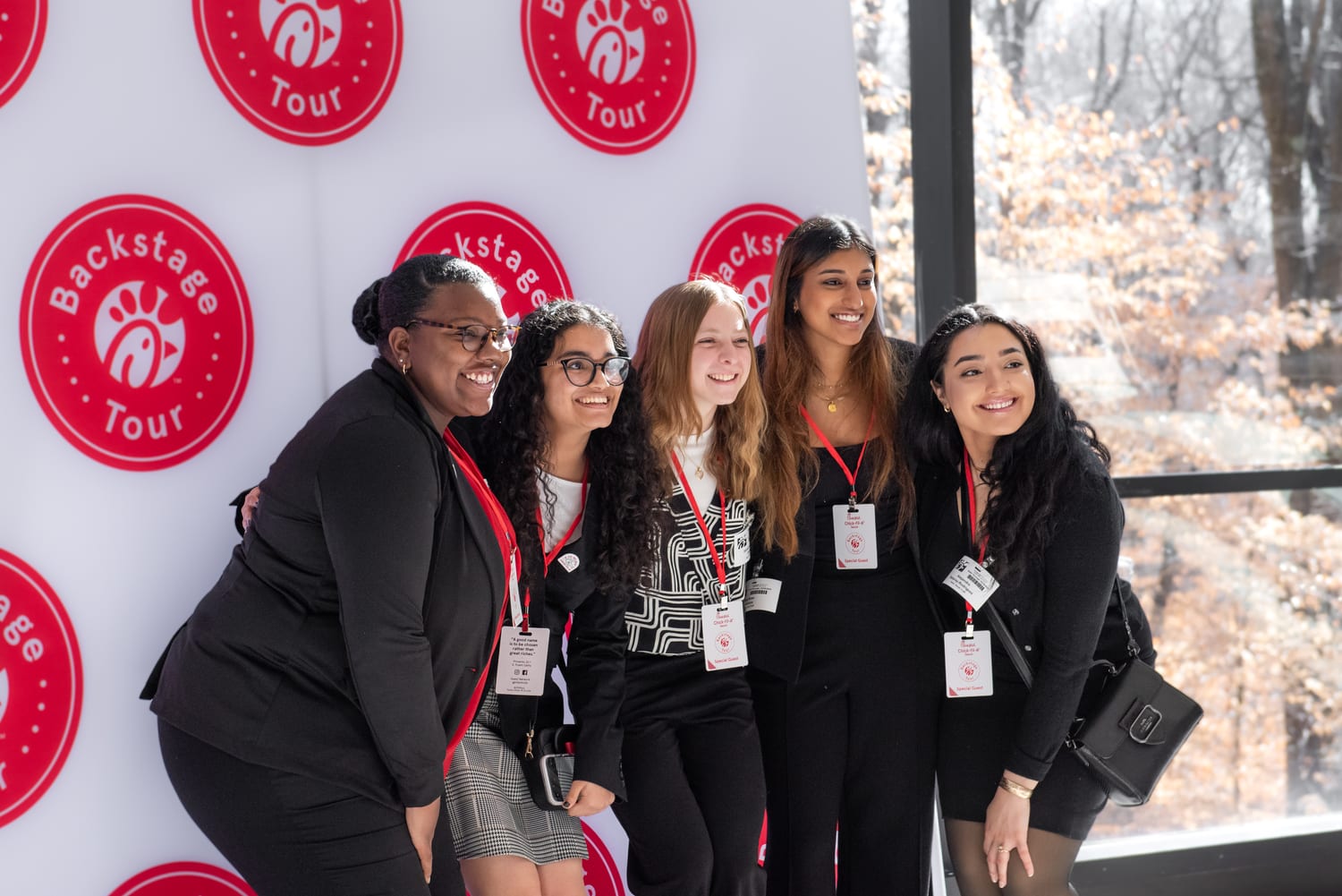 Five scholars in red “Special Guest” lanyards posing in front of a step-and-repeat backdrop tiled with the Chick-fil-A “Backstage Tour” logo, bare winter trees through the window behind them.