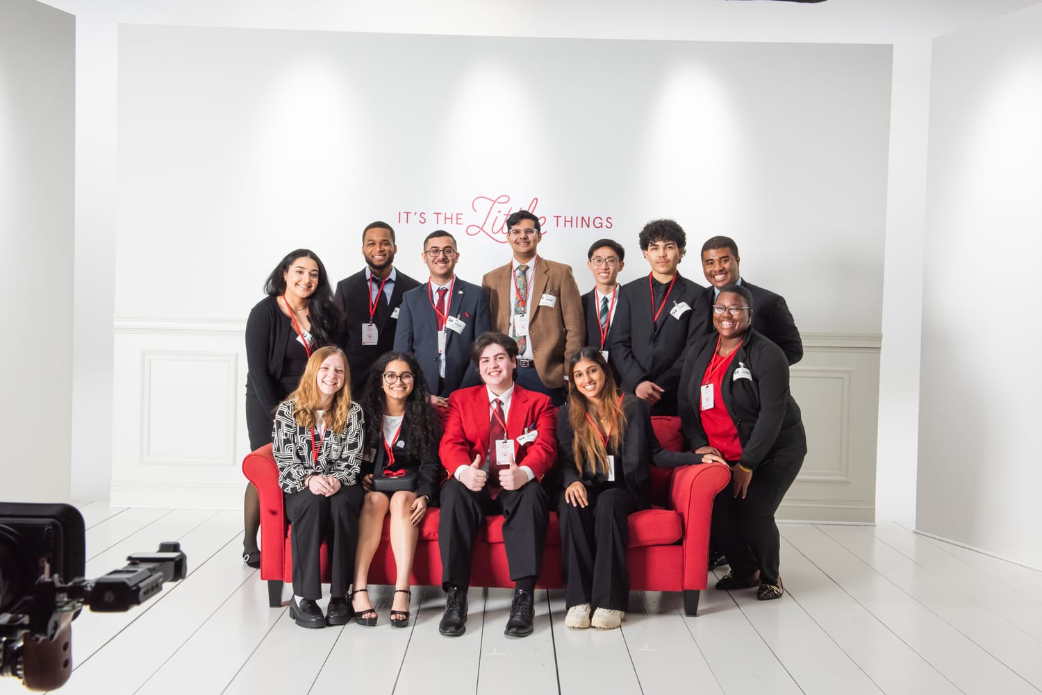 The scholar cohort posed on and around a red loveseat in a white studio, all wearing red lanyards, beneath a wall reading “It’s the Little Things”.