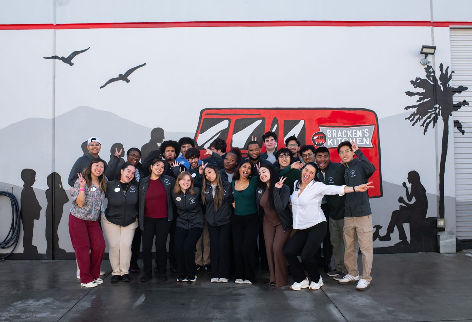 About twenty scholars and staff in matching “Scholars” jackets throwing peace signs in front of a painted mural of a red “Bracken’s Kitchen” food truck.