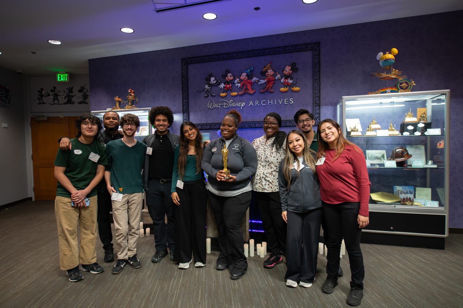 About ten scholars, several in grey “Scholars” jackets, crowded together at the Walt Disney Archives — a Mickey-through-the-ages mural behind them and one scholar holding a gold Oscar statuette.