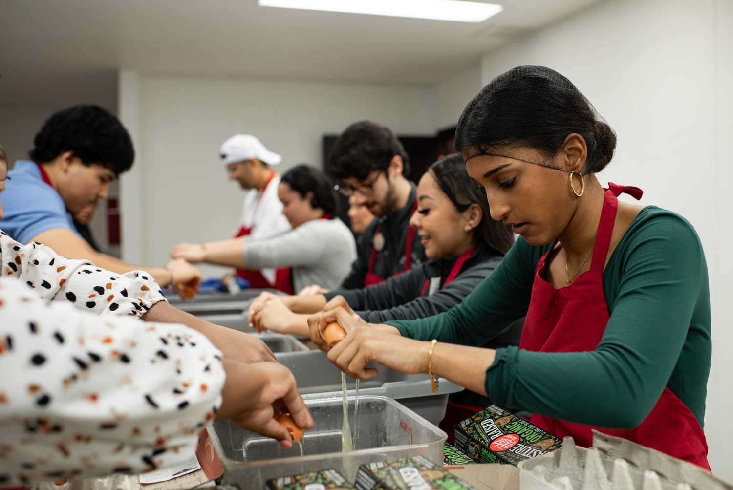 Ava in a hairnet and red apron cracking eggs into a tray alongside a line of fellow scholars prepping food in a community kitchen.