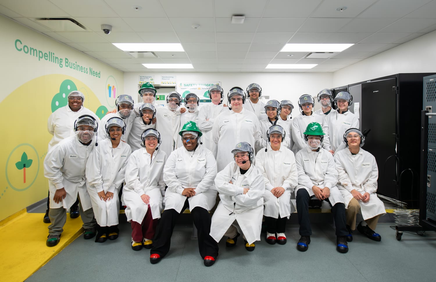 The cohort in white lab coats, hairnets, safety glasses, headsets and hard hats posed inside a food-production facility, a wall behind them reading “Compelling Business Need”.
