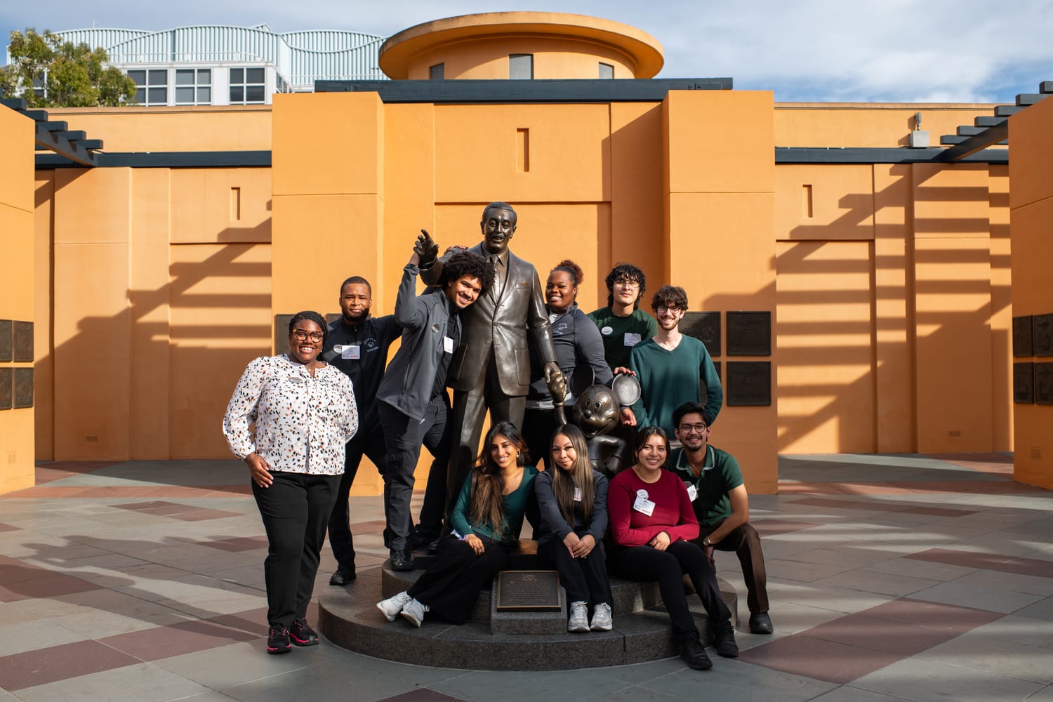 Scholars grouped around the bronze “Partners” statue of Walt Disney and Mickey Mouse in the orange-walled Legends Plaza at the Walt Disney Studios.