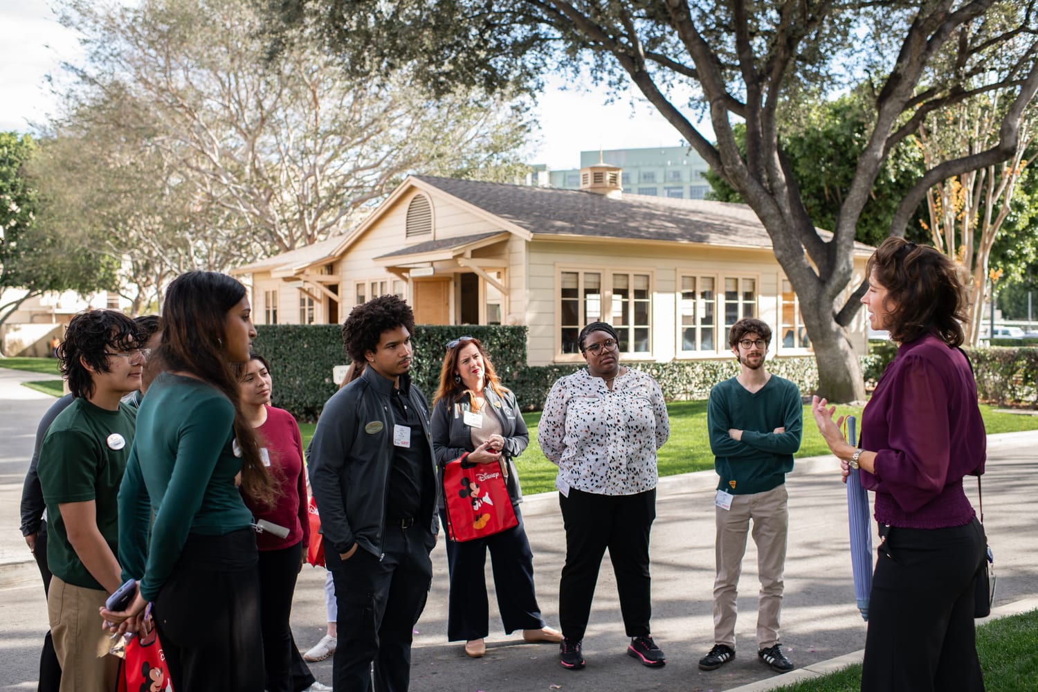 Scholars gathered under a tree on the Walt Disney Studios lot listening to a guide, an original studio bungalow behind them and a red Mickey Mouse tote in hand.