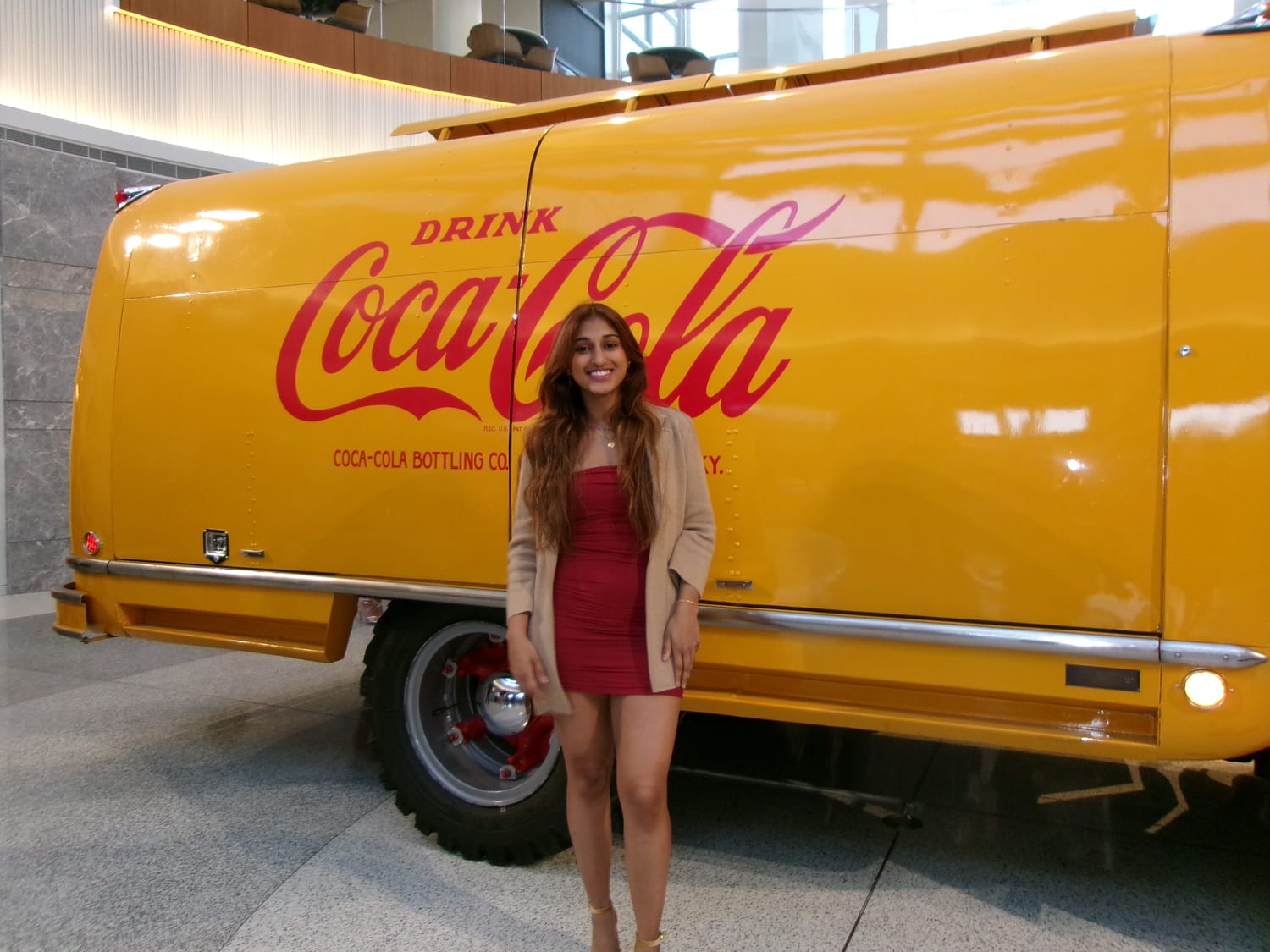 Ava in a red dress and tan cardigan standing beside a restored vintage yellow Coca-Cola delivery truck lettered “Drink Coca-Cola” inside a bright marble atrium.