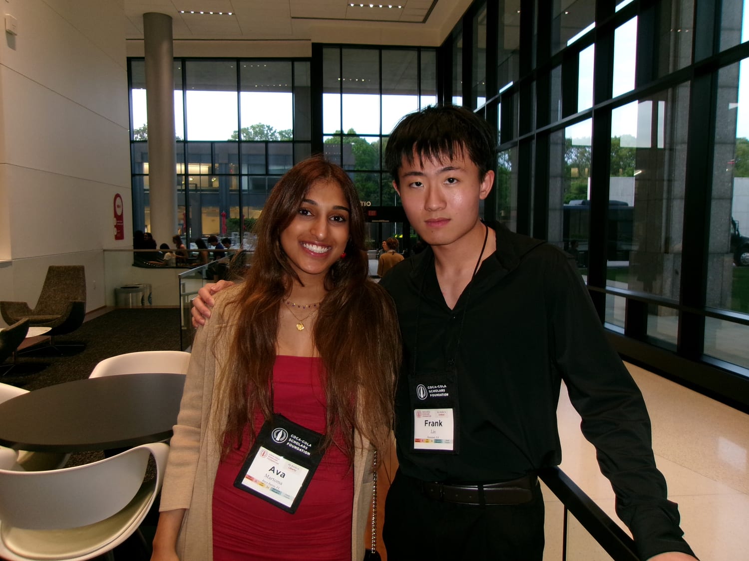 Ava in a red dress, her Coca-Cola Scholars Foundation badge reading “Ava Martoma”, standing arm-in-arm with a fellow scholar in a black shirt in a glass-walled building lobby.