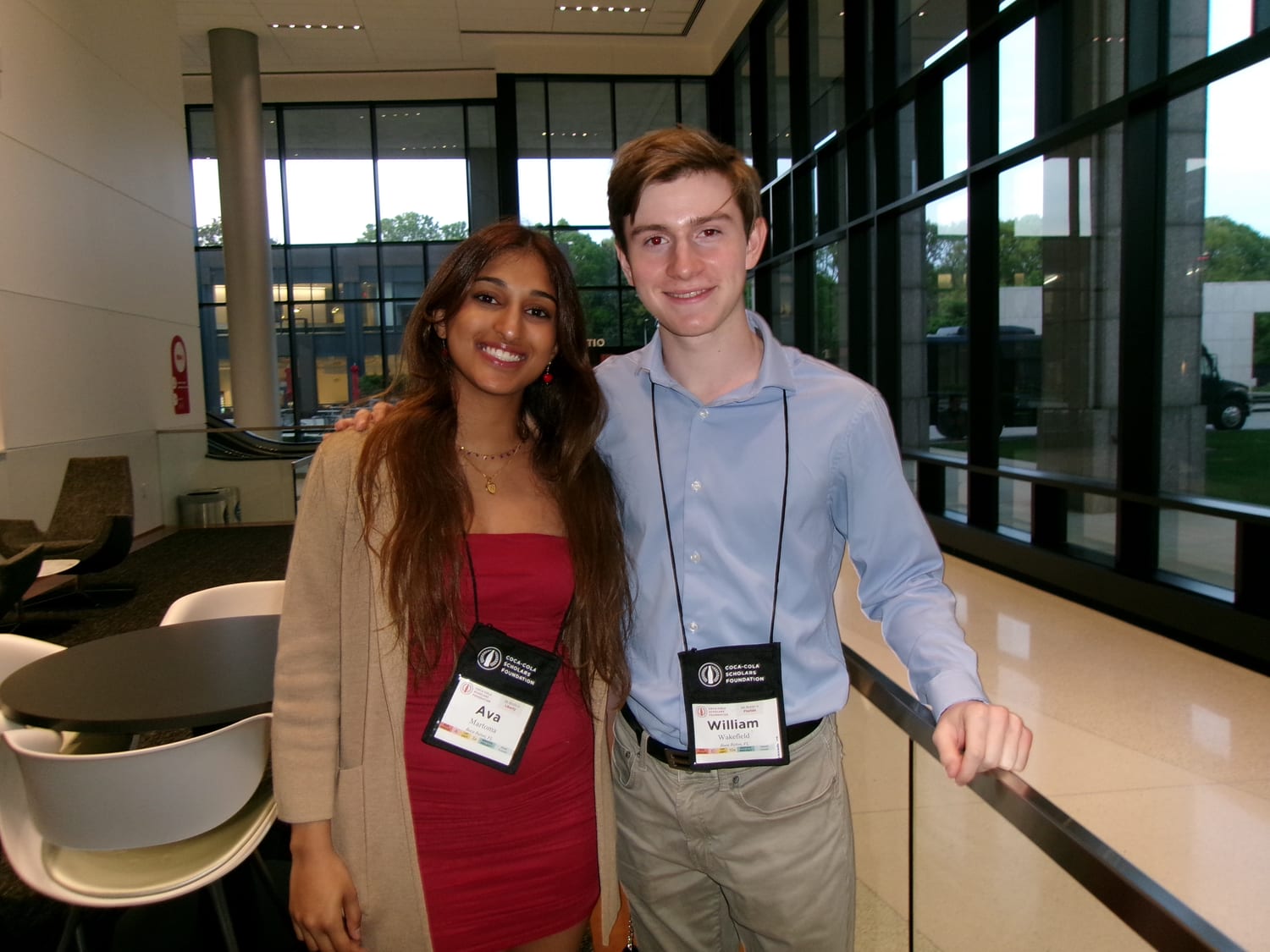 Ava and another Coca-Cola Scholar in a light blue shirt, both wearing Coca-Cola Scholars Foundation lanyards, smiling together by the lobby windows.