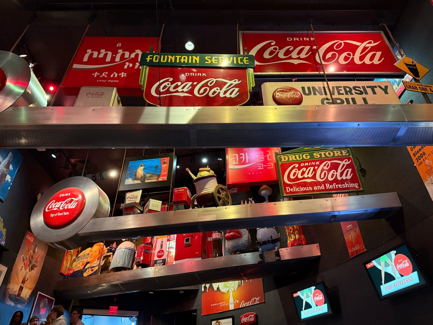 A towering wall of vintage Coca-Cola advertising signs and memorabilia in many languages — “Fountain Service”, “Drug Store — Delicious and Refreshing”, “Sign of Good Taste” — at the World of Coca-Cola.