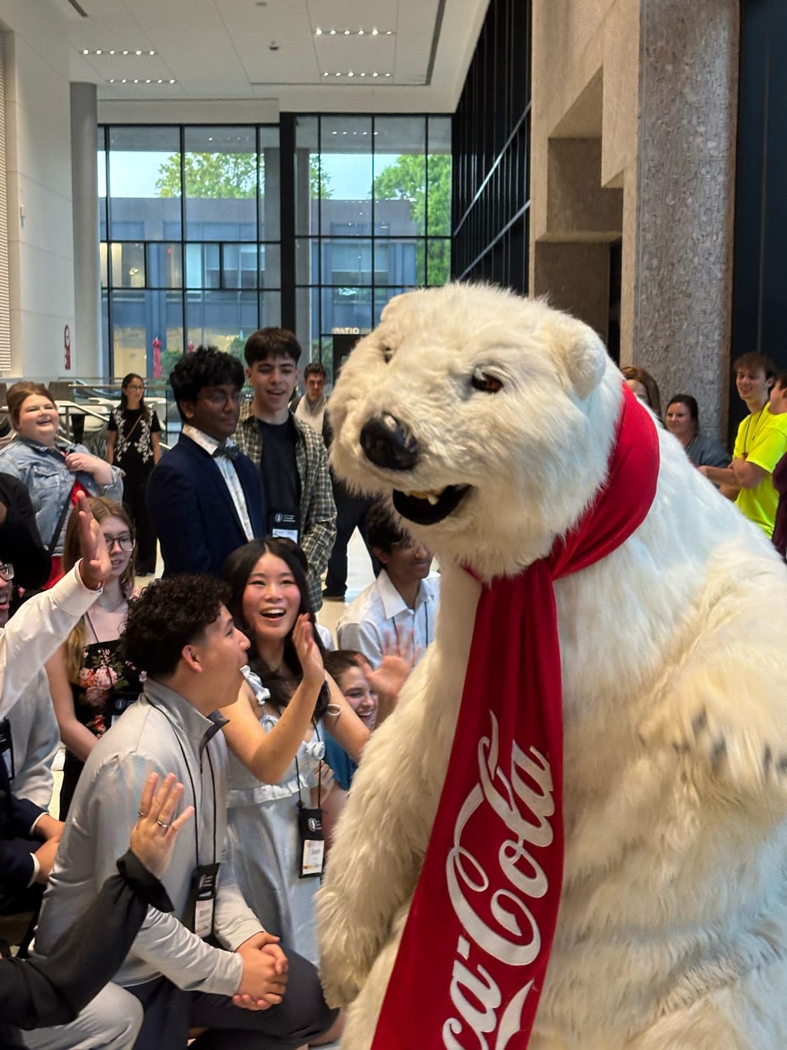 A performer in the Coca-Cola polar bear costume, wearing a red Coca-Cola scarf, greeting a crowd of dressed-up scholars who reach out for high-fives in a glass-walled lobby.