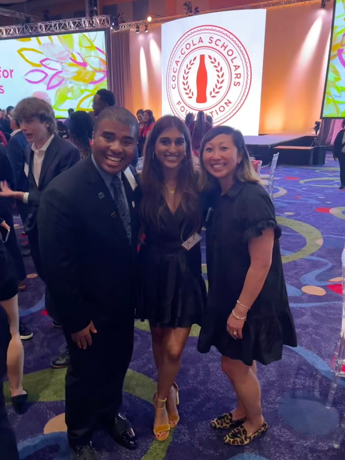 Ava in a black dress standing between two people at the banquet, the round Coca-Cola Scholars Foundation logo lit up on the backdrop behind them and a colorful patterned carpet underfoot.