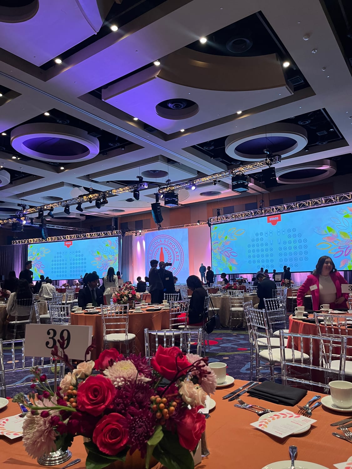 The banquet hall set for the gala — round tables in orange linens with silver chiavari chairs and red rose centerpieces, twin stage screens carrying the Coca-Cola Scholars Foundation seal.