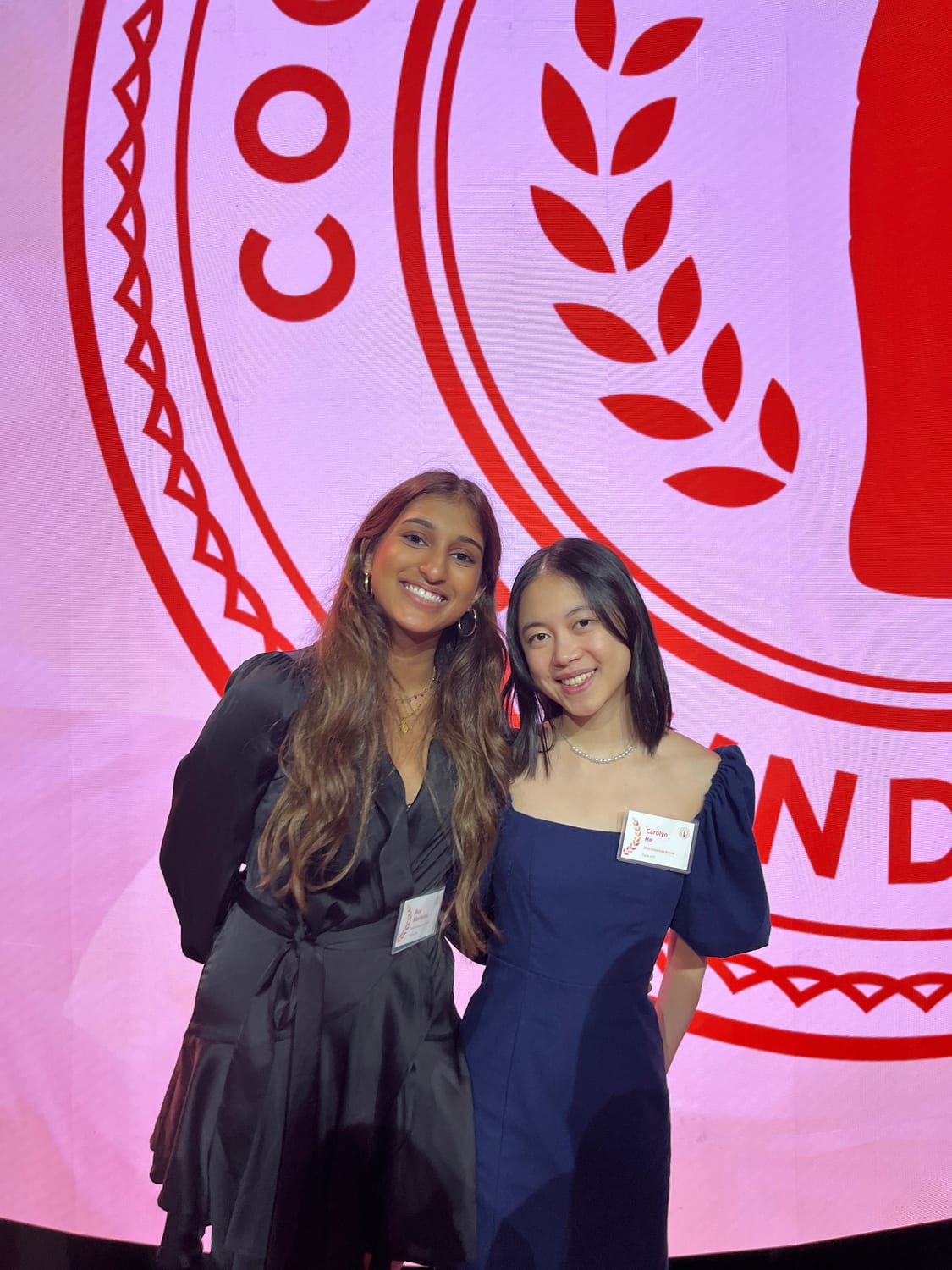 Ava in a black dress with a fellow scholar in navy in front of an enormous illuminated red Coca-Cola Scholars seal glowing pink and red.