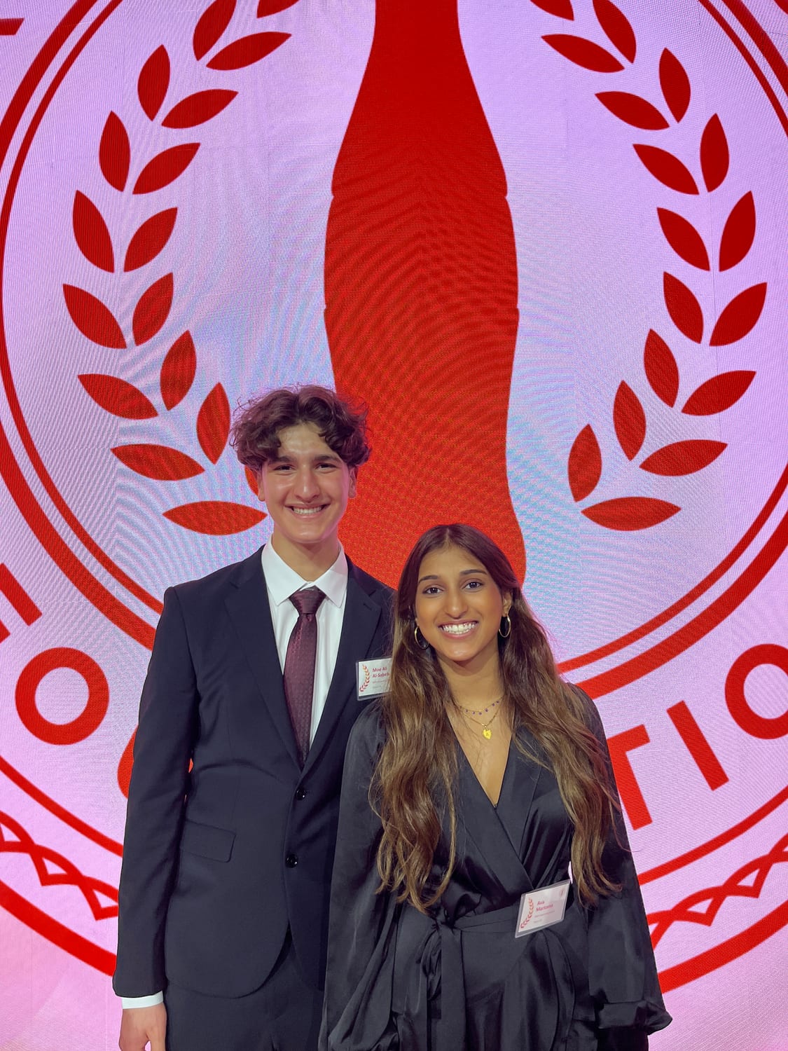 Ava in a black dress standing with a fellow scholar in a dark suit in front of the giant red Coca-Cola bottle-and-laurel seal backdrop.