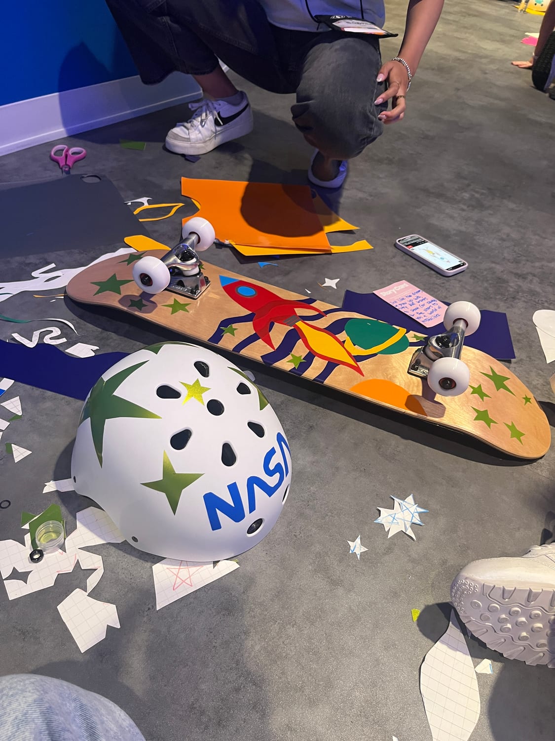 A service-project craft in progress on the floor: a wooden skateboard deck decorated with a cut-paper rocket and stars, and a white helmet marked “NASA”, surrounded by paper scraps.