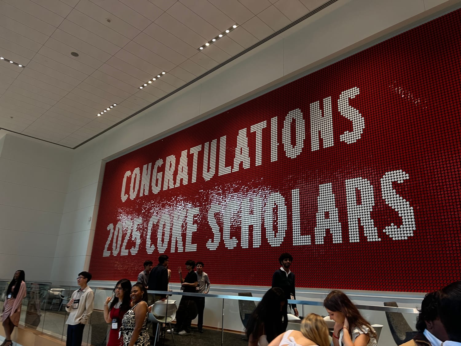 A giant red-and-white peg-art wall spelling “Congratulations 2025 Coke Scholars”, with scholars gathered on the balcony below it.