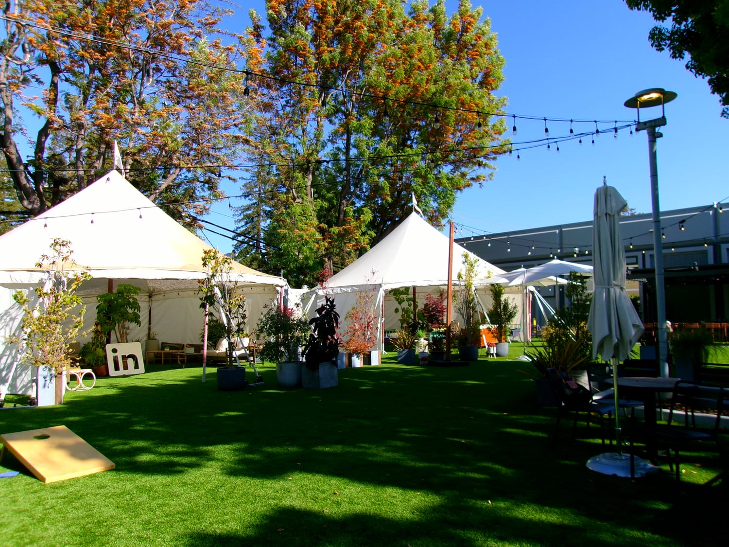 A LinkedIn campus lawn set up for an event: white peaked tents strung with festoon lights over artificial turf, a freestanding white “in” logo sign, a cornhole board, and patio tables under a bright blue sky.