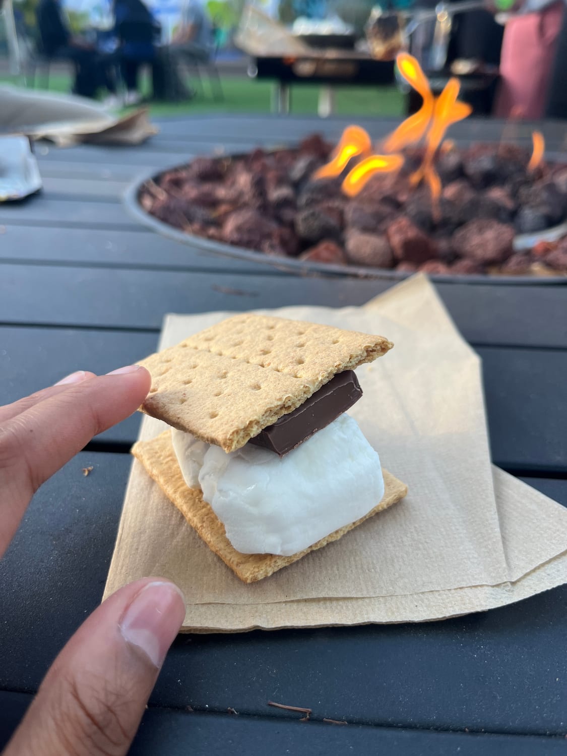 A hand holding an assembled s’more — toasted marshmallow and chocolate between graham crackers — on a napkin in front of a lit gas fire pit at dusk.