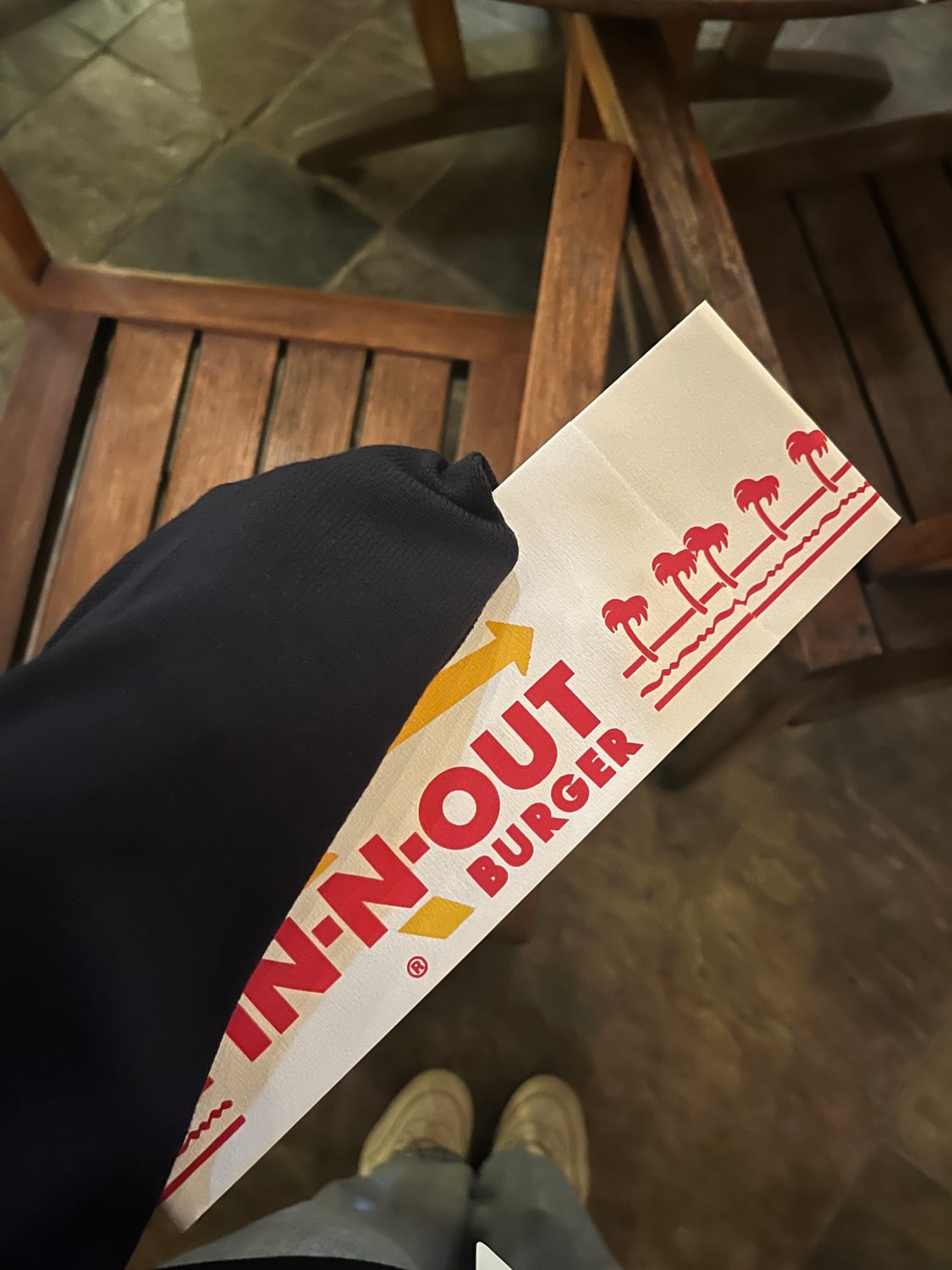 Point-of-view shot of a folded paper In-N-Out Burger hat resting on a lap, wooden patio chairs and sneakers below.