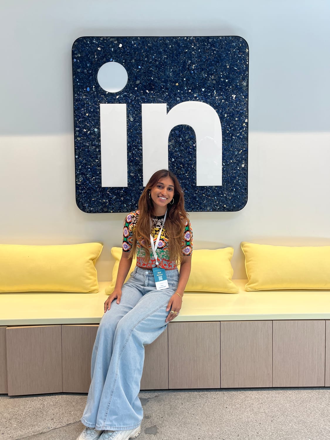 Ava in a colorful floral top and wide-leg jeans, wearing a teal LinkedIn lanyard, sitting on a yellow bench beneath a large dark-blue glittered LinkedIn “in” logo mounted on the wall.