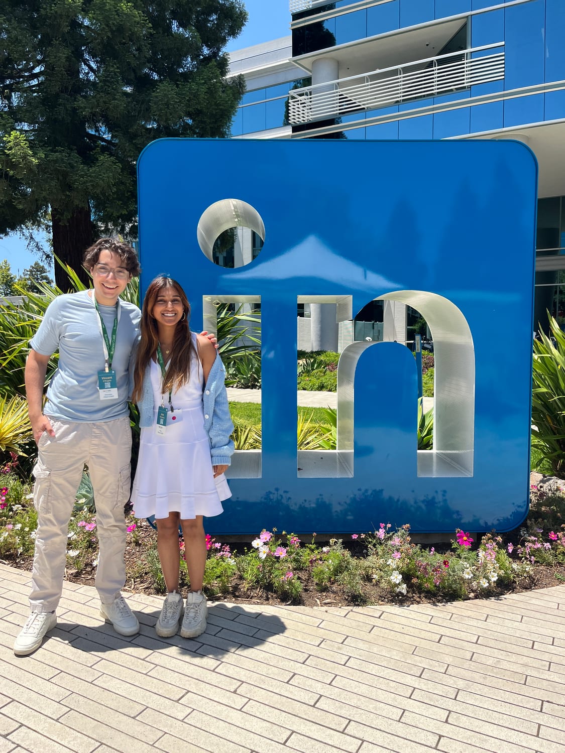 Ava in a white dress and blue cardigan standing with a fellow scholar in front of the giant glossy blue LinkedIn “in” sculpture outside on the campus, a flowerbed at their feet.