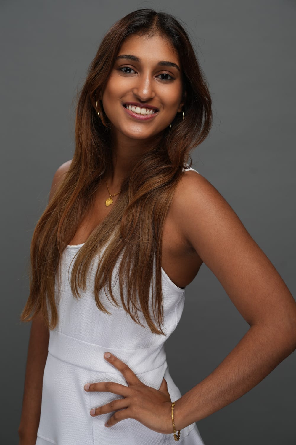 A studio headshot of Ava in a white dress against a grey backdrop, smiling with one hand on her hip.