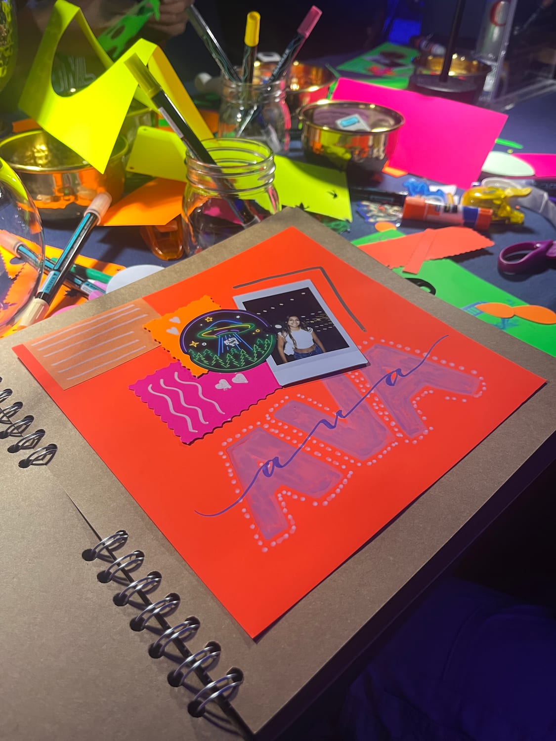A scrapbook page under blacklight: a neon-orange sheet with “AVA” painted in bubble letters, a polaroid, a glowing UFO sticker and decorative washi tape, on a table scattered with neon craft supplies.