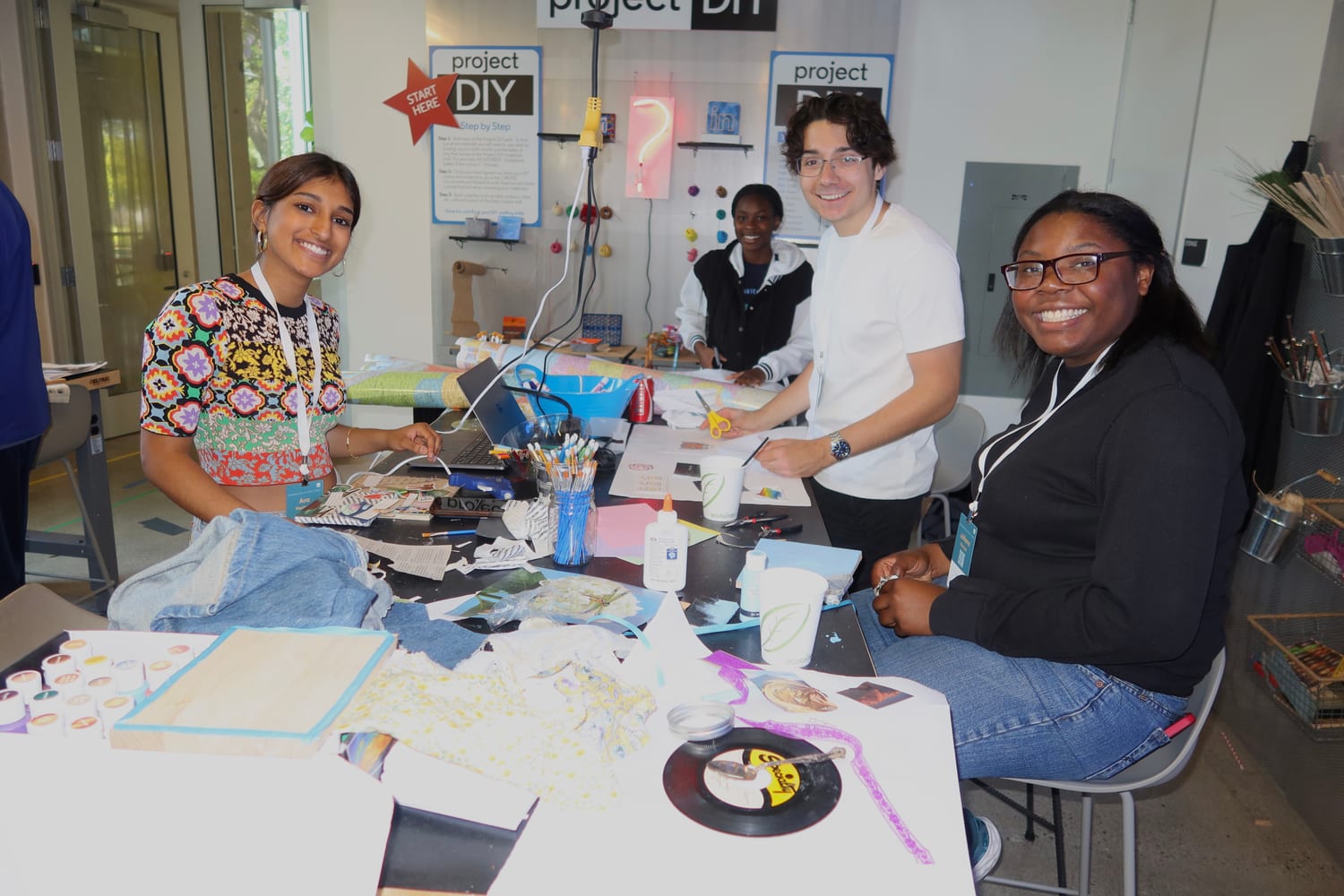 Ava and three other scholars smiling around a “project DIY” maker-station table strewn with paper, glue, brushes, and collage materials.