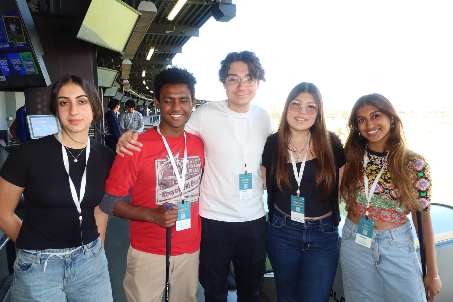 Ava and four other scholars in lanyards posing arm-in-arm at a Topgolf bay, one holding a golf club, screens and driving-range netting behind them.