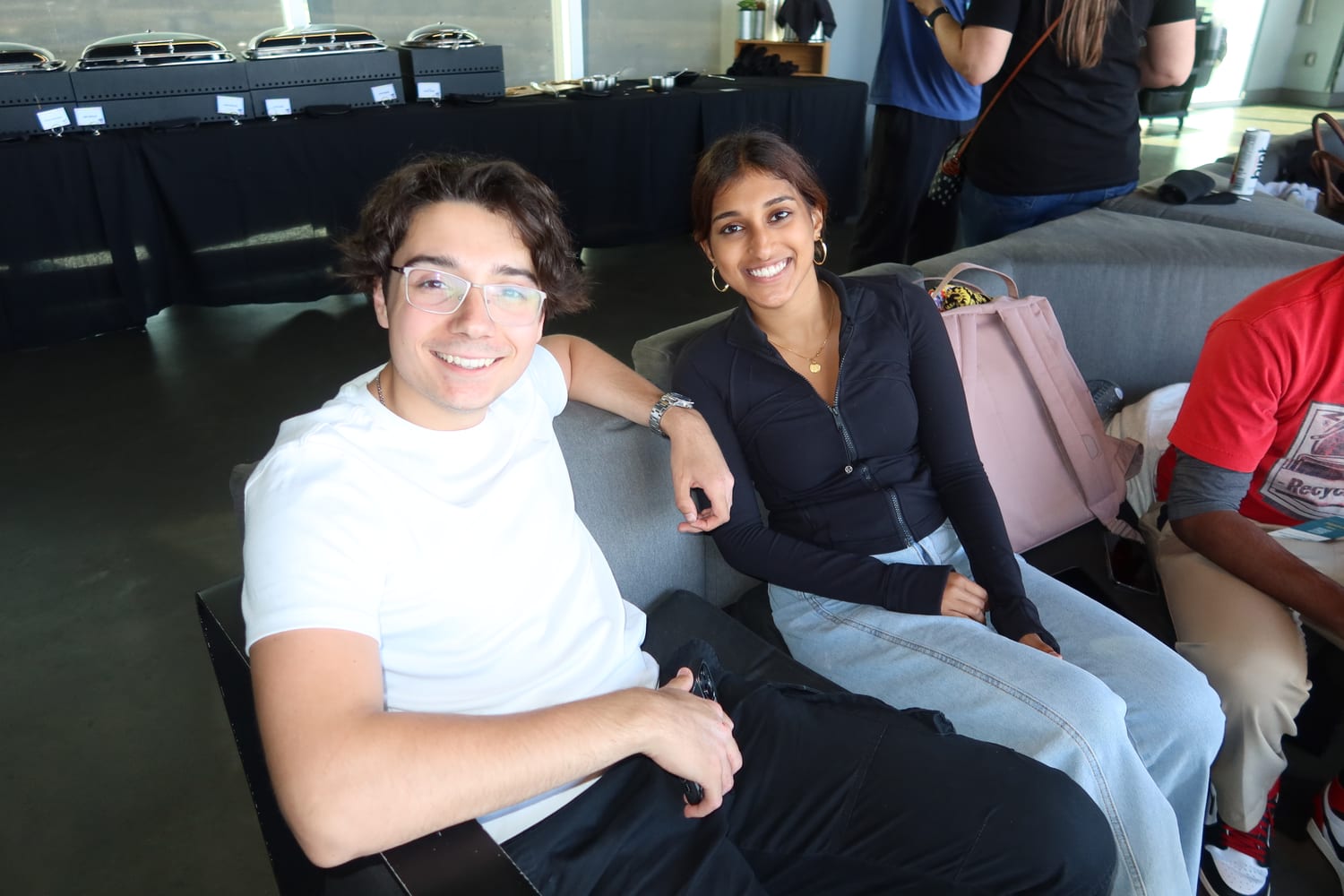 Ava and a fellow scholar sitting side by side on grey lounge chairs, smiling, with catering chafing dishes on a table behind them.