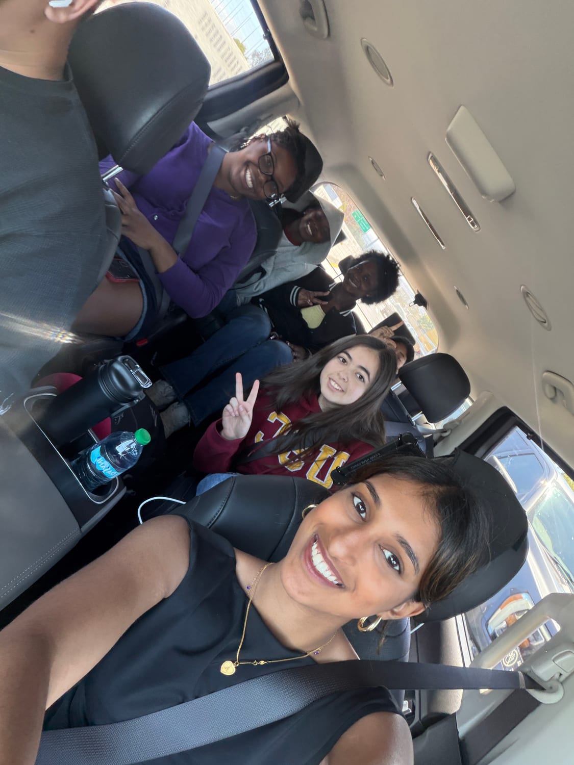 A tilted selfie inside a van packed with scholars heading to an activity, one flashing a peace sign and Ava smiling in the front seat with her seatbelt on.