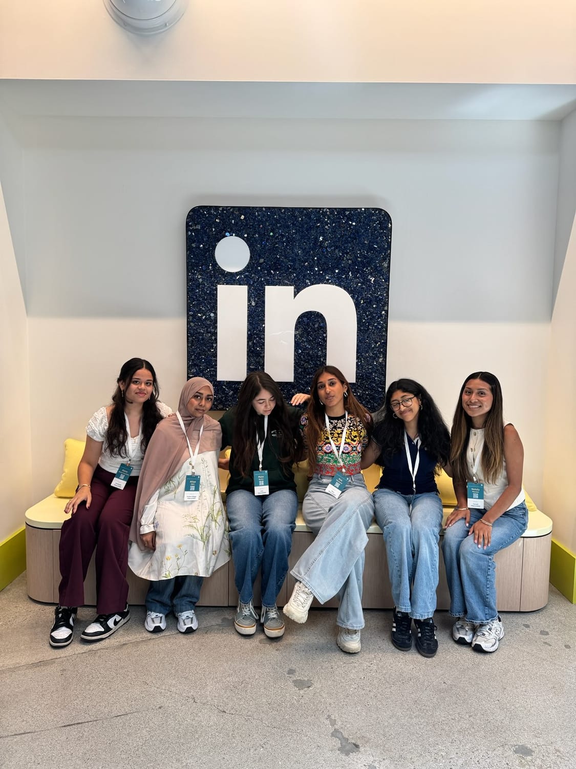 Six scholars, including Ava, sitting shoulder to shoulder on a yellow bench beneath the glittered blue LinkedIn “in” logo indoors.