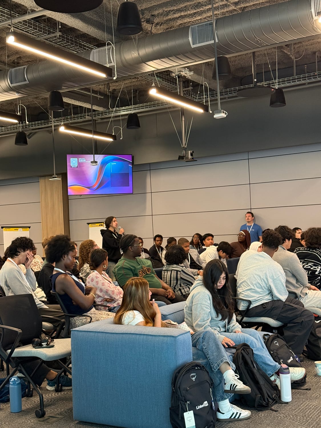 A student stands with a microphone asking a question during a session, seated scholars filling the lounge chairs around her and a LinkedIn-branded backpack on the floor in front.