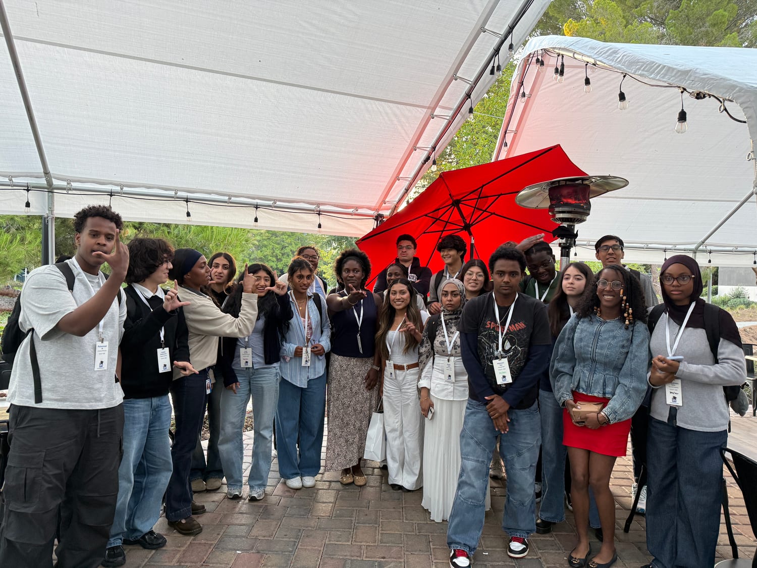 About twenty scholars crowded together under a white event tent at dusk, a red patio umbrella and outdoor heater behind them, several throwing shakas and finger-hearts.