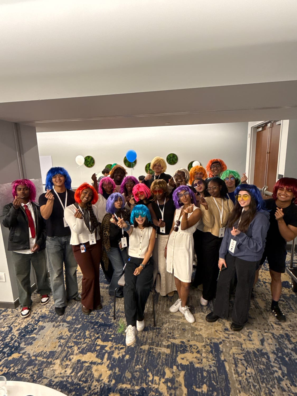About twenty scholars in brightly colored costume wigs — pink, blue, purple, orange, green — crowded together throwing finger-hearts, balloons on the wall behind them.