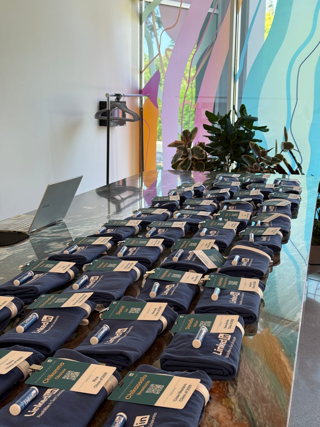 A long marble table lined with rows of folded navy LinkedIn “Possibilities in Tech Scholarship” T-shirts, each paired with a green name tag listing a scholar’s name and college “Class of 2029” and a branded lip balm.