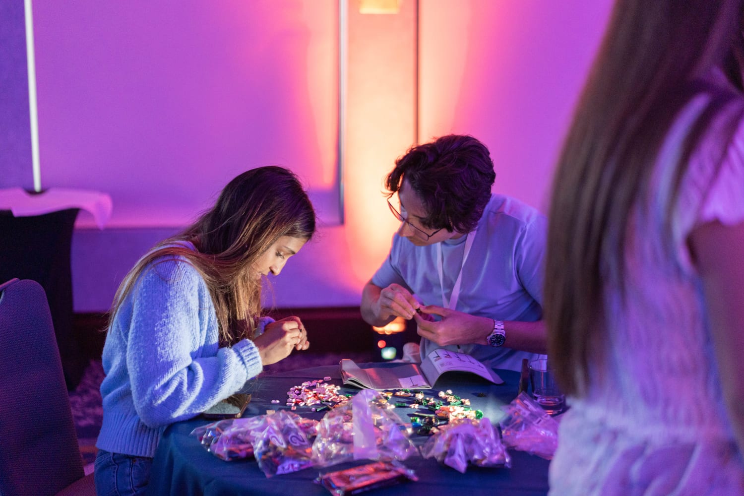 Ava and a fellow scholar bent over a table making beaded crafts under purple and orange mood lighting, bags of beads and charms spread out in front of them.