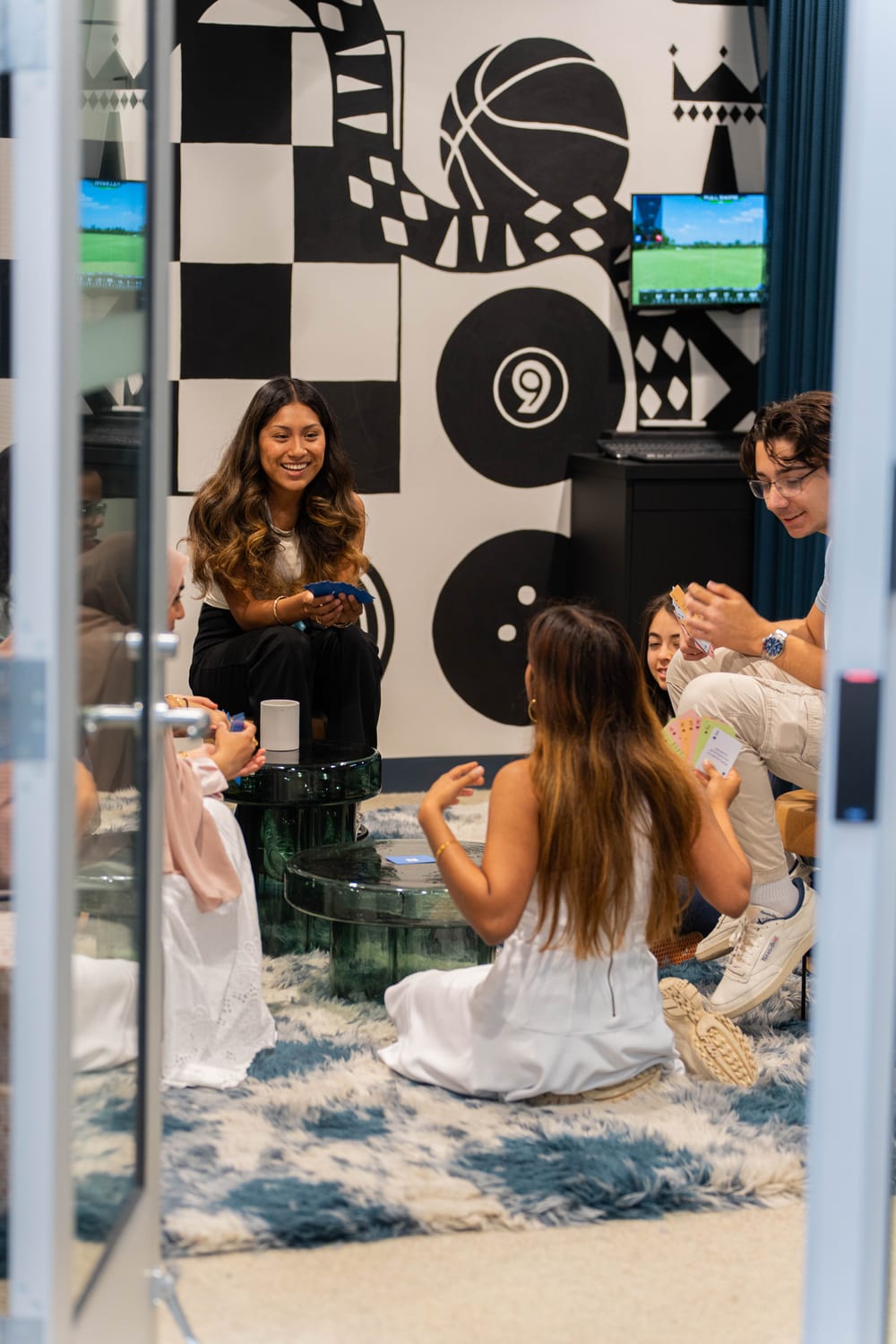 Seen through a doorway, a group of scholars sit on the floor and stools around glass tables playing a card game in a game room with a bold black-and-white sports mural.