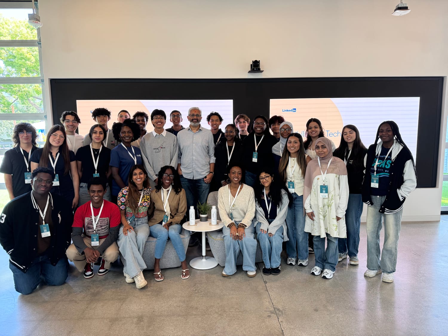 The full scholarship cohort — about thirty students in lanyards — grouped around a LinkedIn host on a couch in a bright, window-lined space, LinkedIn screens glowing behind them.