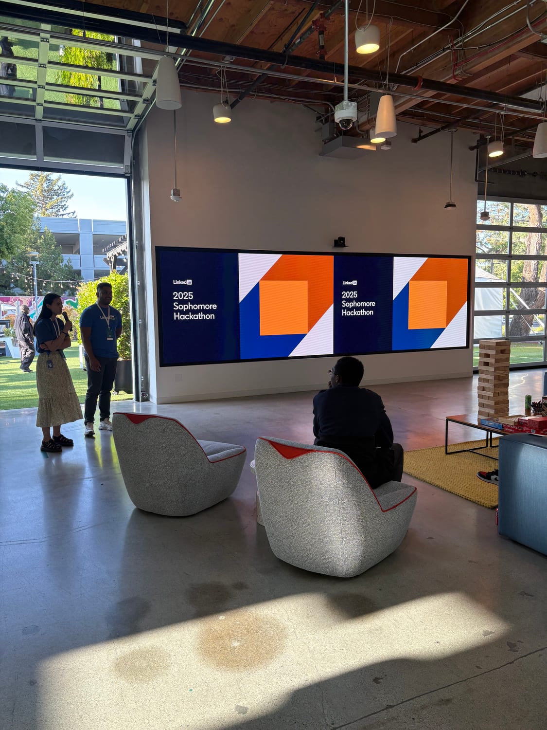 A LinkedIn loft space with two wall screens reading “LinkedIn — 2025 Sophomore Hackathon”, a presenter with a microphone by the open garage door, lounge chairs and a Jenga tower on the floor.