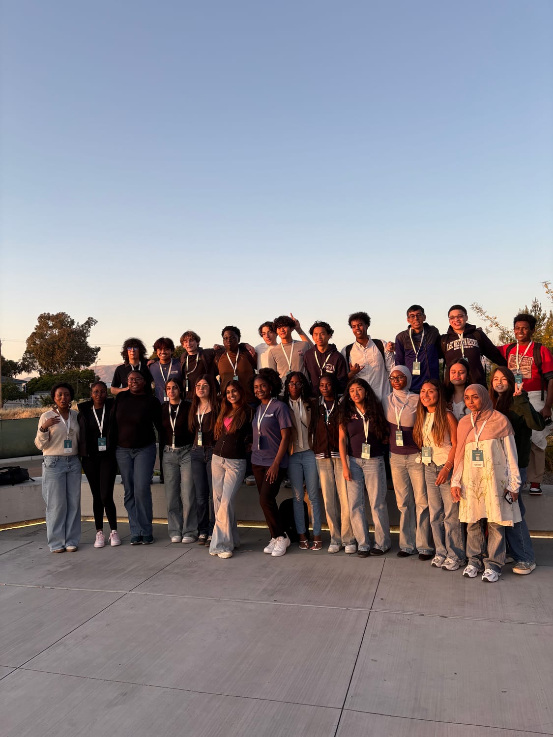 The cohort gathered in two rows on an outdoor concrete plaza at golden hour under a clear sky, arms around each other for a group portrait.