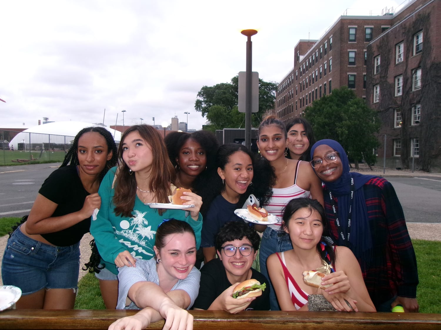 Ten students crowded around a picnic table outside an MIT dorm holding burgers and hot dogs at a cookout.