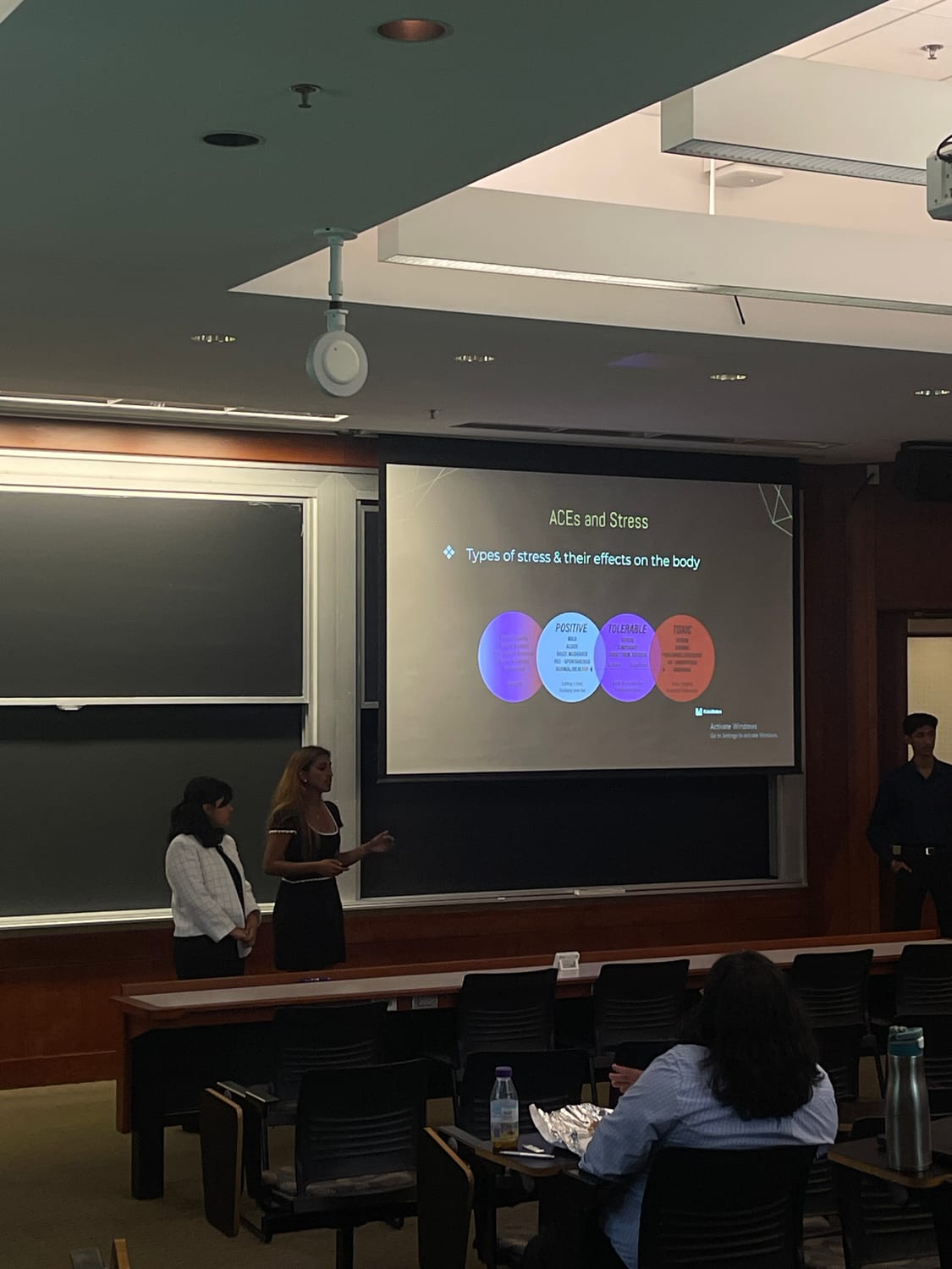 Ava presenting in a lecture hall beside a slide titled “ACEs and Stress — types of stress and their effects on the body”, carrying the KidsMates logo.