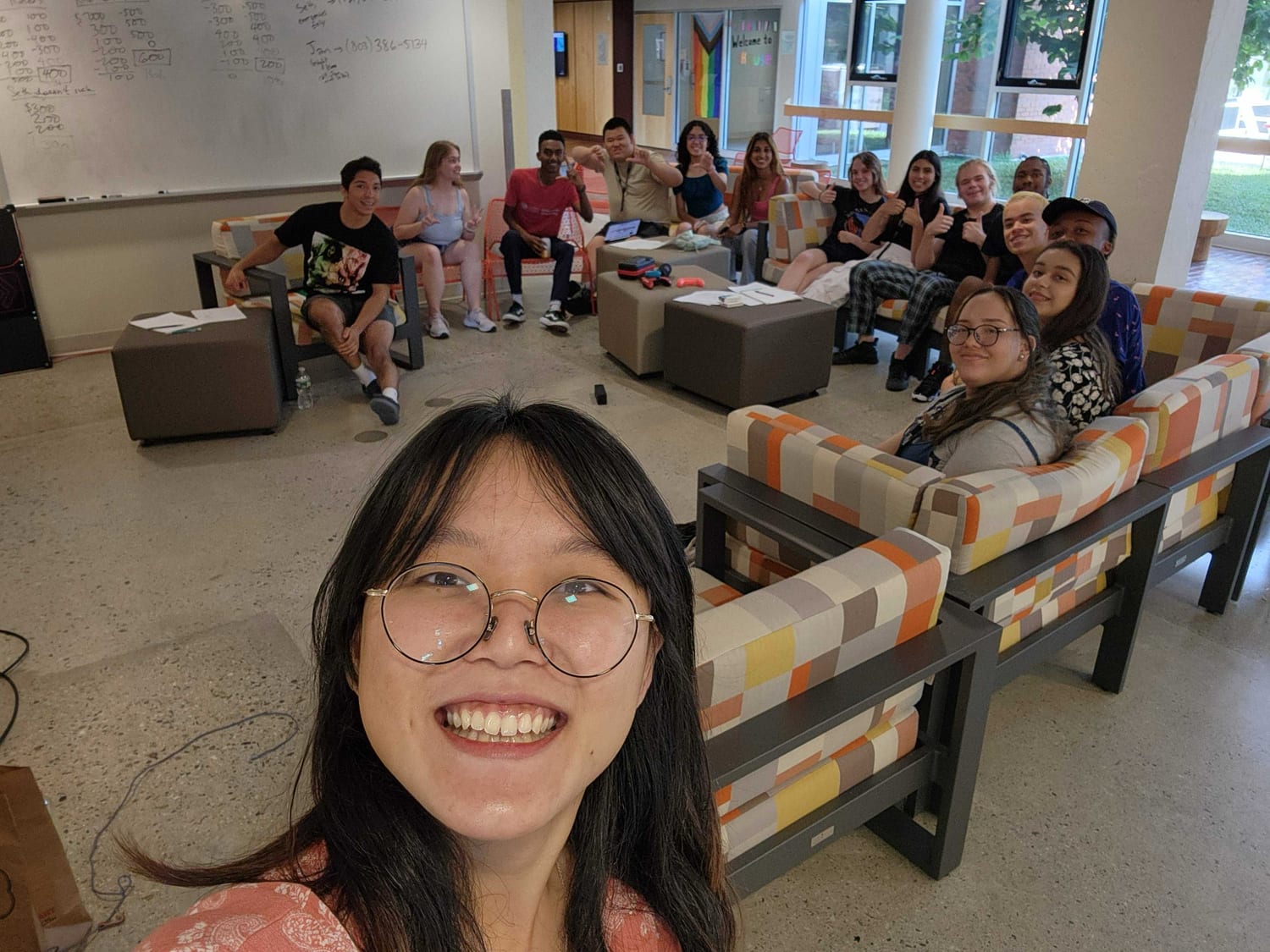 A selfie of a dozen students spread across the couches of a common room, giving thumbs up, a whiteboard of figures behind them.