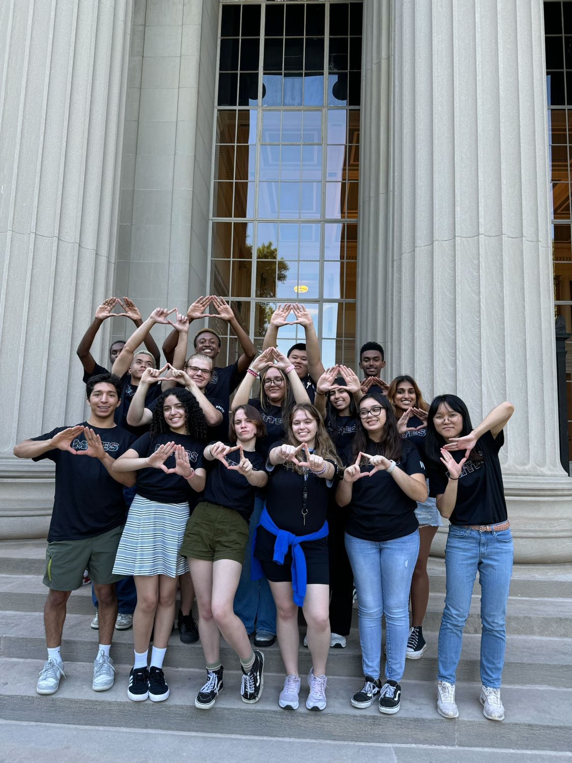 The MITES cohort in matching black MITES T-shirts on the columned steps of an MIT building, all making the pyramid hand sign.